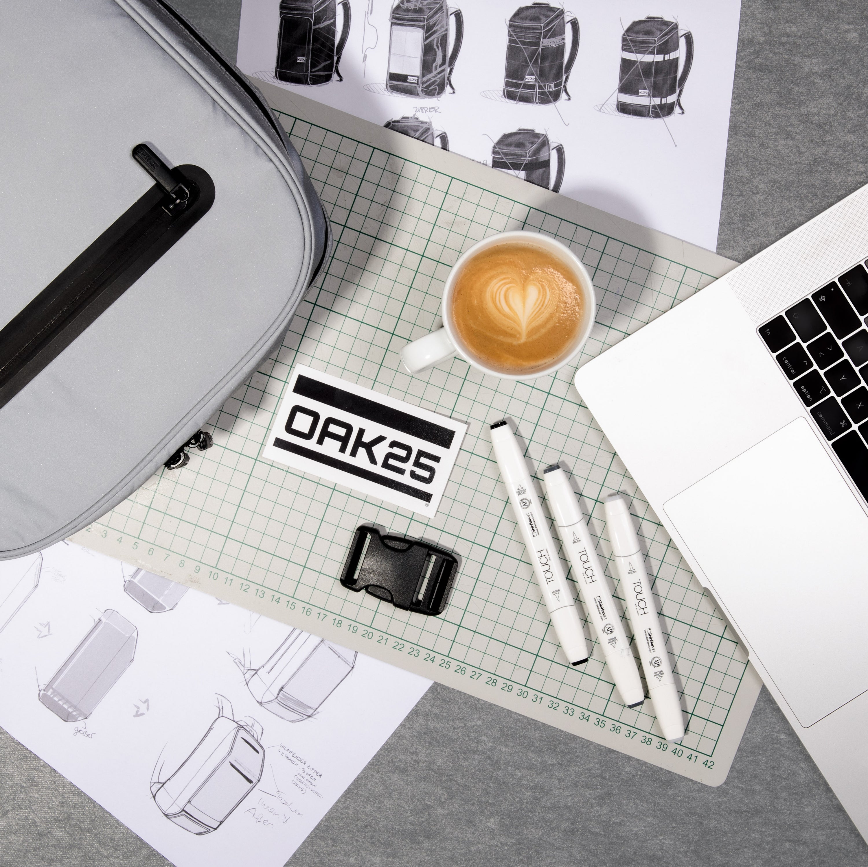 Arbeitsplatz mit grauem Rucksack, Laptop, weißen Markern, Kaffeetasse mit Latte Art, Entwurfsskizzen von Rucksäcken und OAK25 Etikett