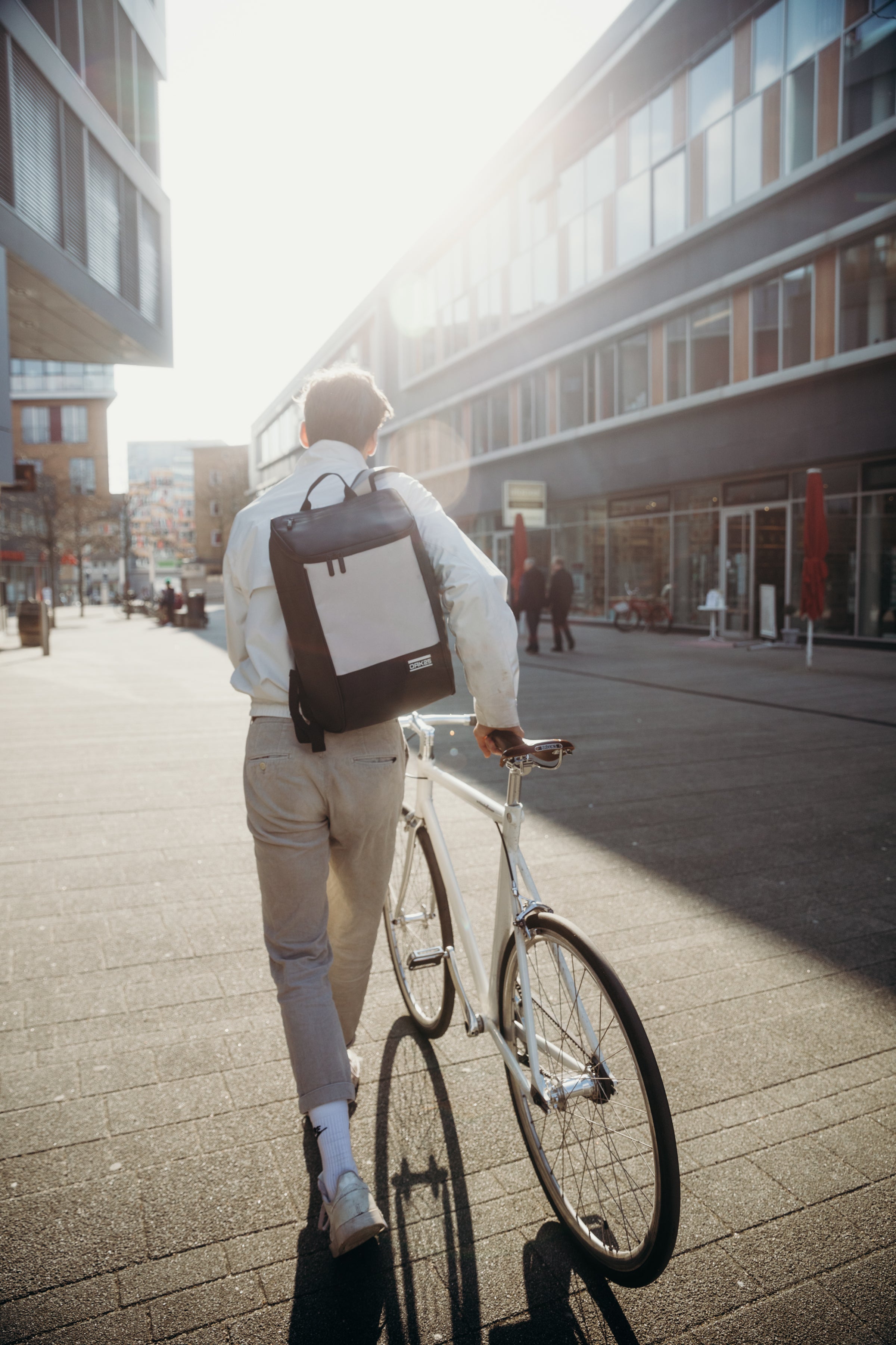 Person mit weißer Jacke und schwarzem Rucksack schiebt weißes Fahrrad auf städtischem Gehweg bei Sonnenlicht
