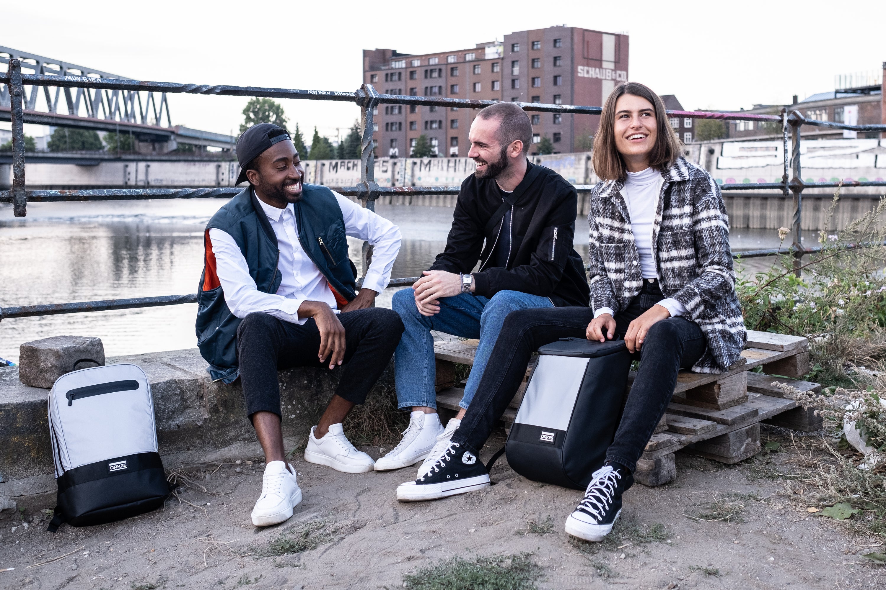 Drei lächelnde junge Erwachsene sitzen an einem Flussufer, zwei mit grauschwarzen Rucksäcken