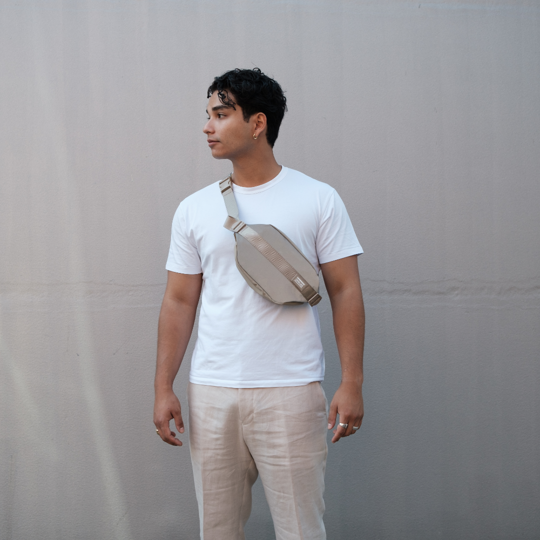 Junger Mann trägt eine sandfarbene, reflektierende Crossbody-Tasche über weißem T-Shirt vor grauer Wand