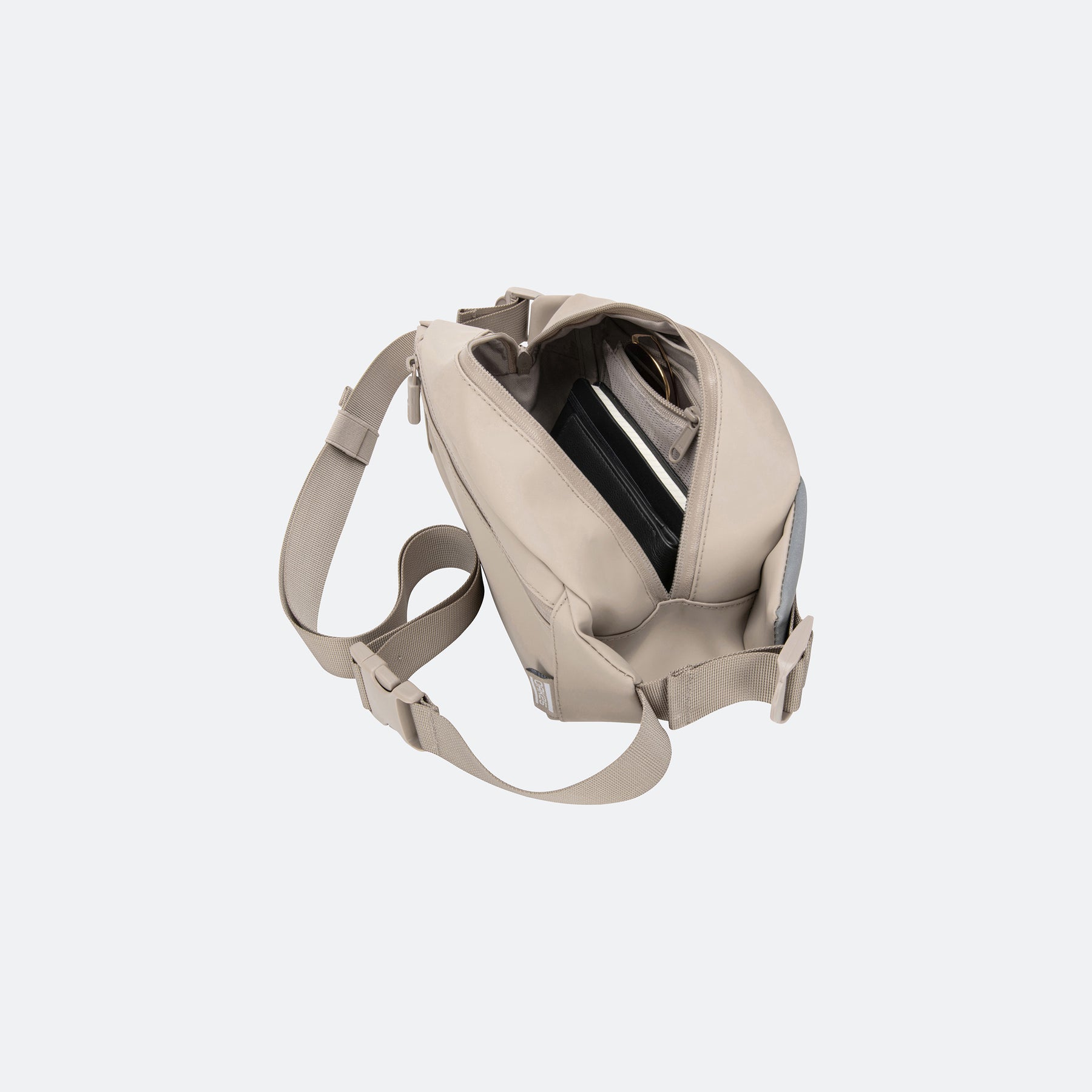 Sandfarbene Sling-Tasche mit geöffnetem Hauptfach, darin ein Notizbuch und eine Sonnenbrille