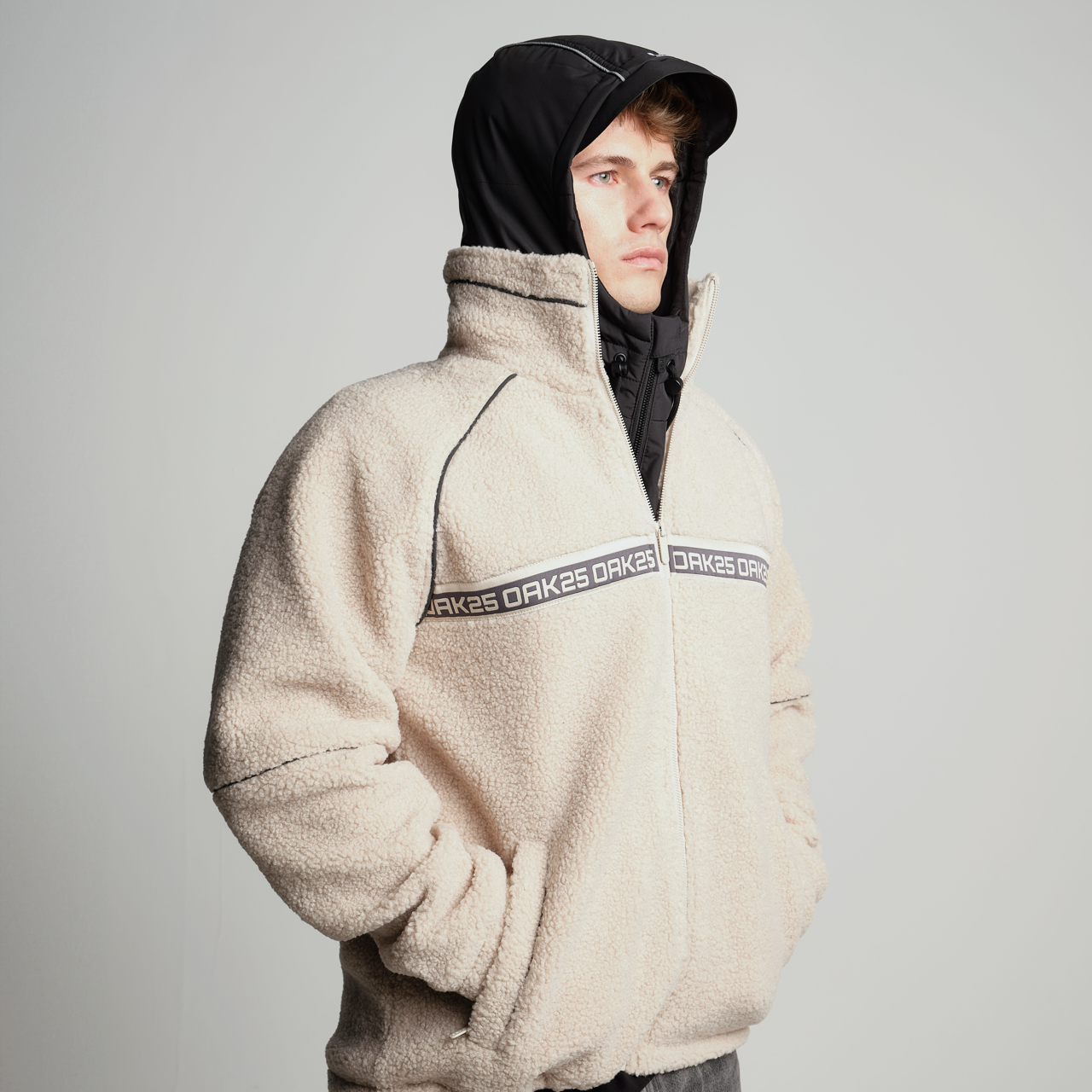 Mann mit schwarzem Hoodie unter beiger Fleecjacke mit 'OAK25'-Logo auf Brusthöhe