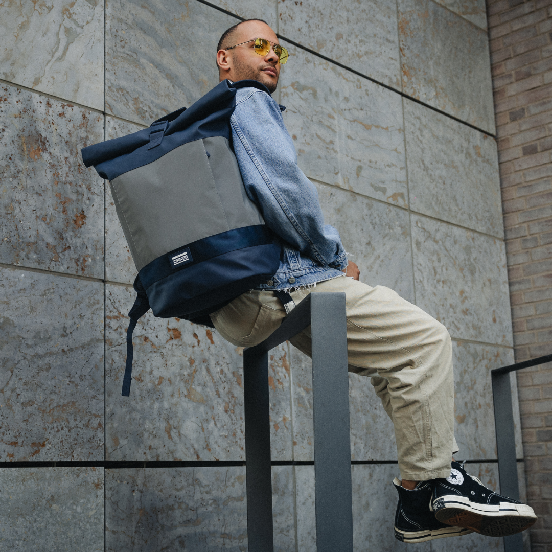 Mann mit gelber Brille und Jeansjacke trägt einen zweifarbigen, navy-grauen Rolltop-Rucksack von OAK25 und sitzt auf einem grauen Geländer vor einer Steinwand