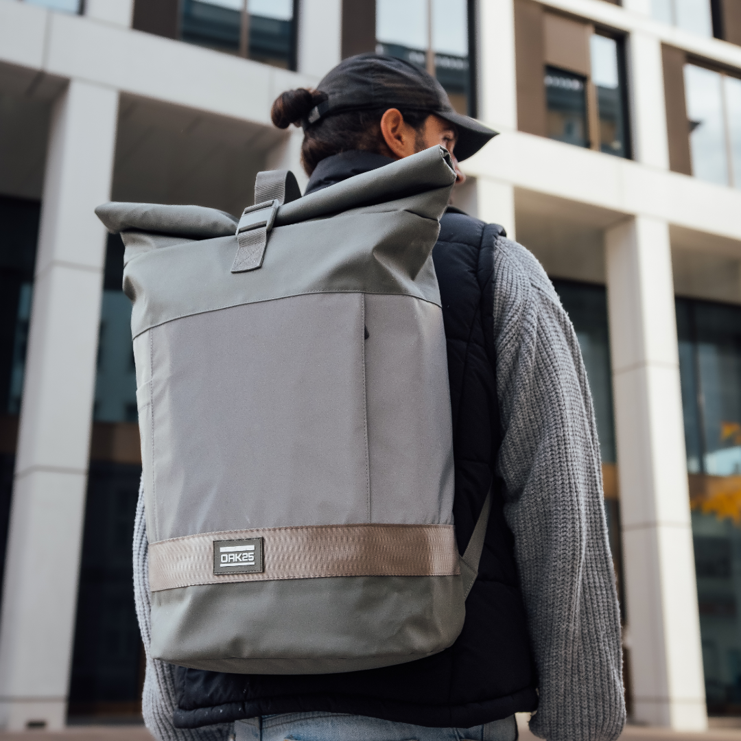 Salbeigrüner Rolltop-Rucksack mit beigem Riemen und OAK25-Logo, getragen von Person in urbaner Umgebung
