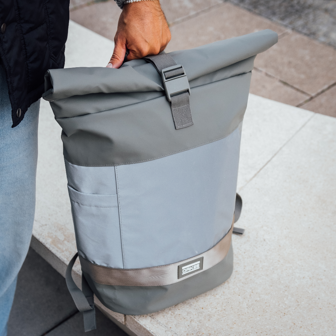 Männliche Hand hält salbei- und graufarbigen Rolltop-Fahrradrucksack mit reflektierendem Streifen