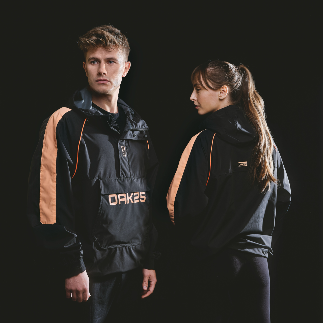 Mann und Frau tragen schwarzen unisex Windbreaker mit pfirsichfarbenen Streifen an den Ärmeln, vorne und hinten mit OAK25-Logo