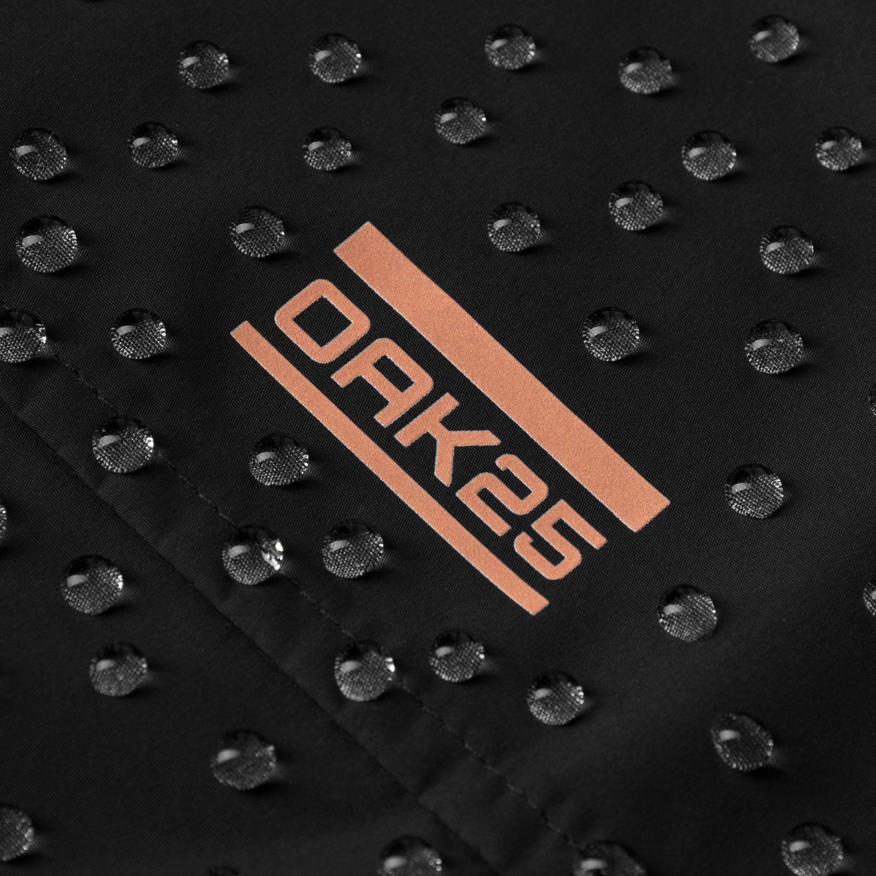Wassertropfen auf schwarzem, wasserabweisendem Stoff mit orangem 'OAK25' Logo