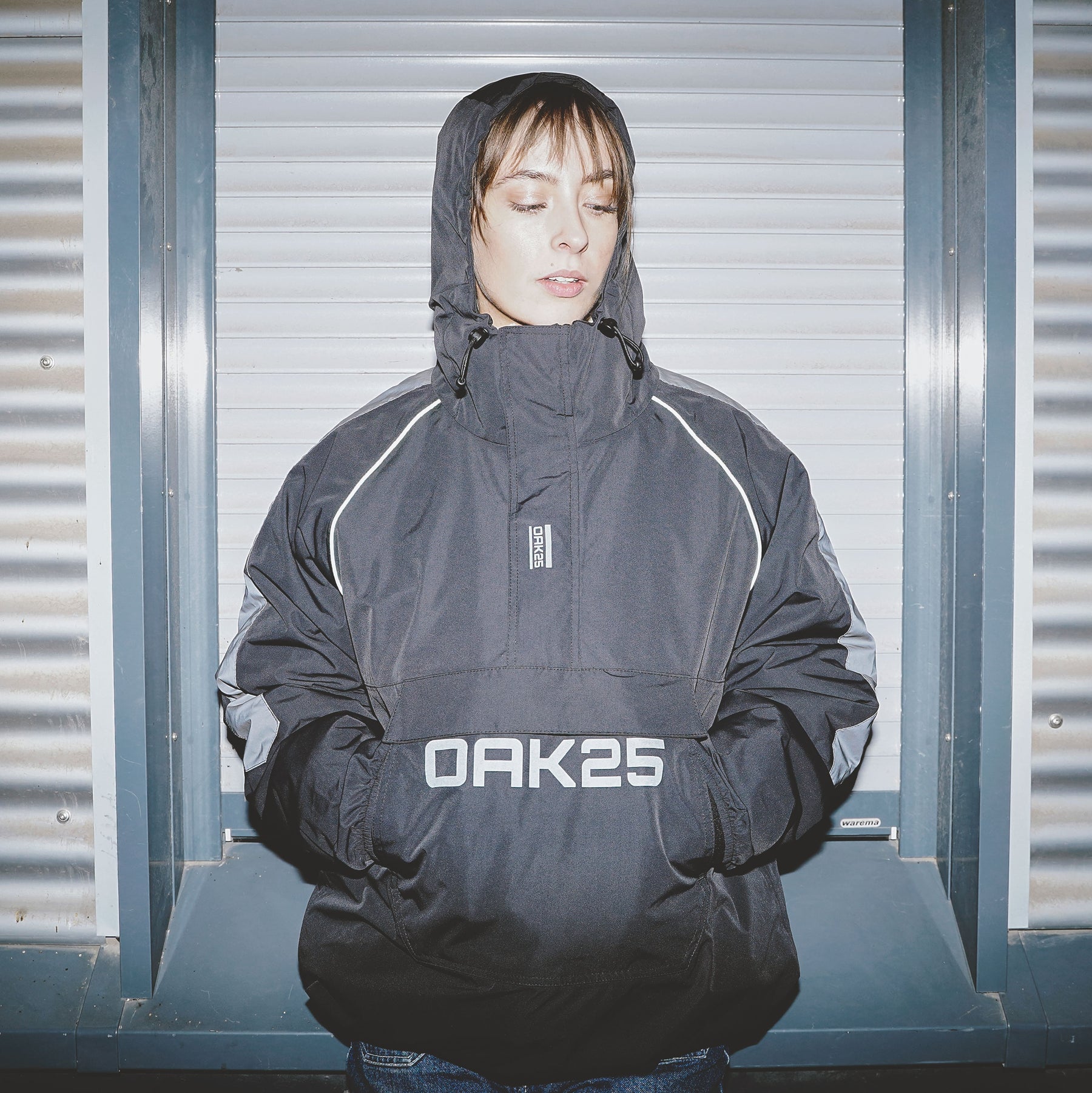 Frau trägt schwarzen OAK25 Windbreaker mit Kapuze und reflektierenden Details vor Metallladen
