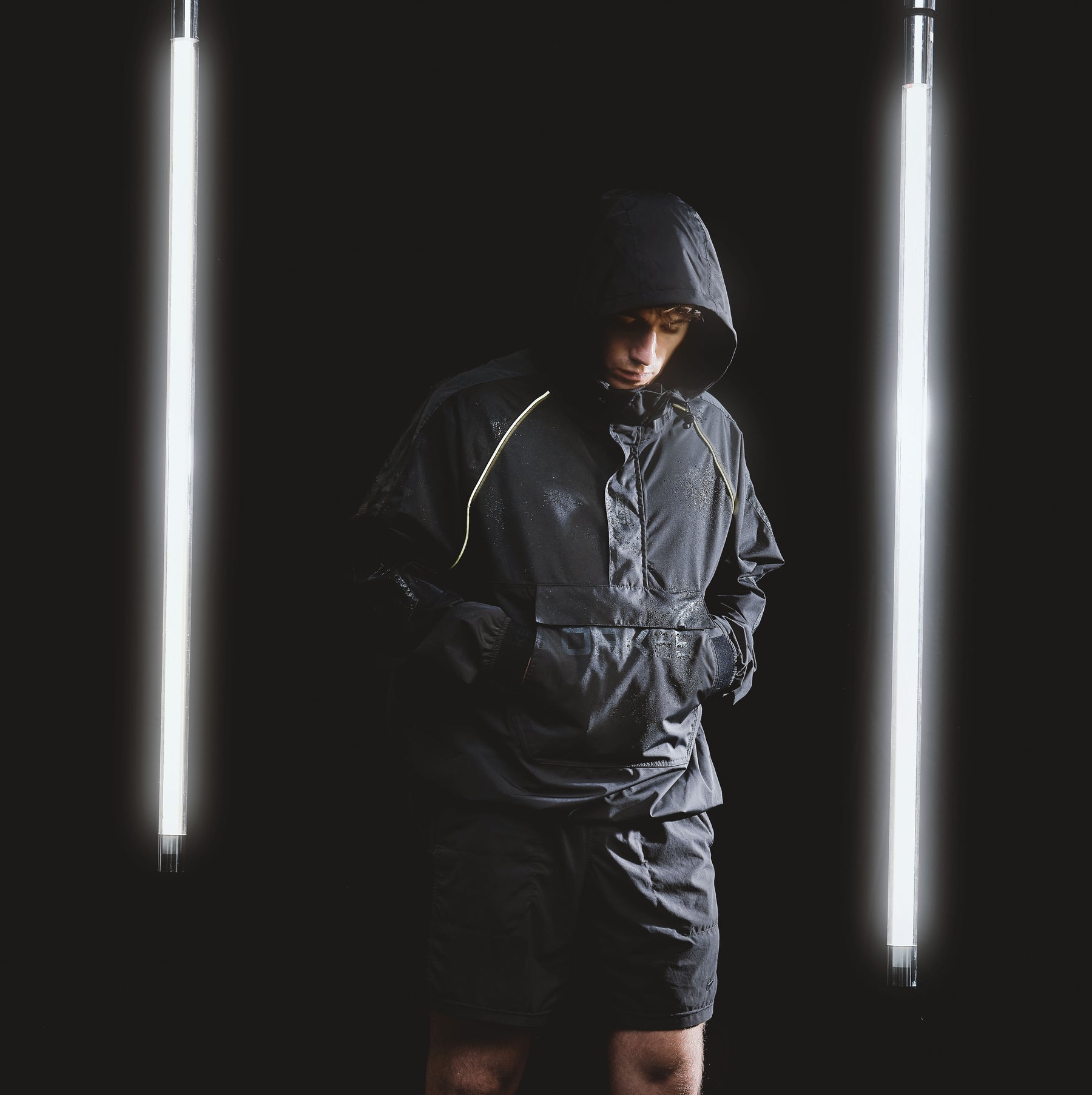 Mann trägt schwarzen, regendichten Windbreaker mit Kapuze und Shorts, umgeben von zwei vertikalen Leuchtstoffröhren