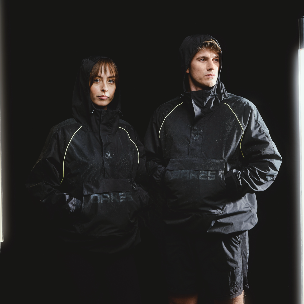 Zwei Personen tragen schwarze, wasserfeste Windbreaker mit Kapuze und reflektierenden Details, unisex Modell von OAK25