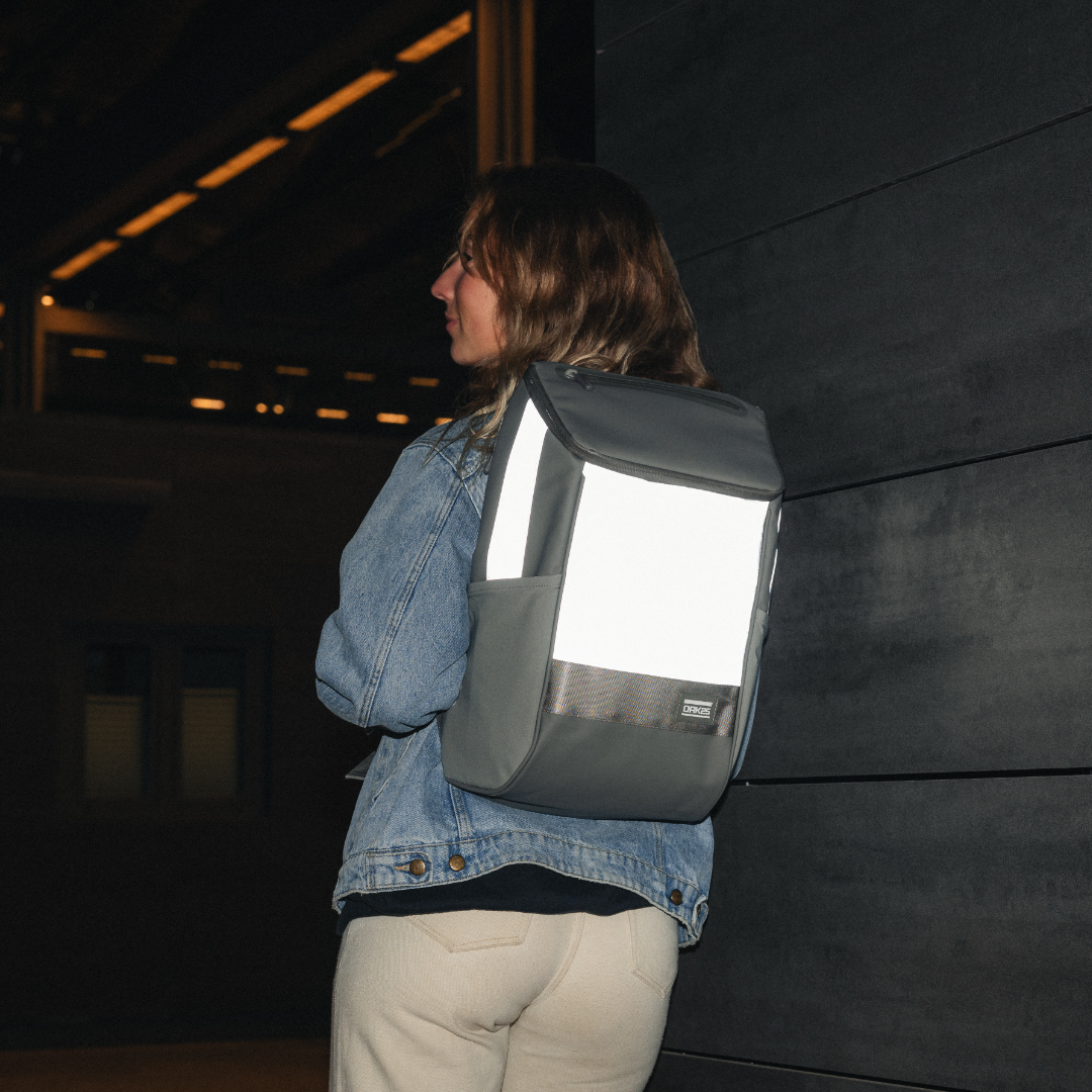Frau mit reflektierendem, salbeigrünem Daybag-Rucksack an einer dunklen Wand