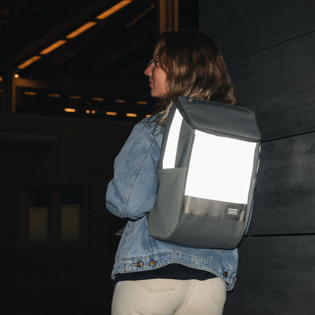 Frau trägt reflektierenden Daybag-Rucksack in Salbeigrün bei Nacht vor dunkler Wand