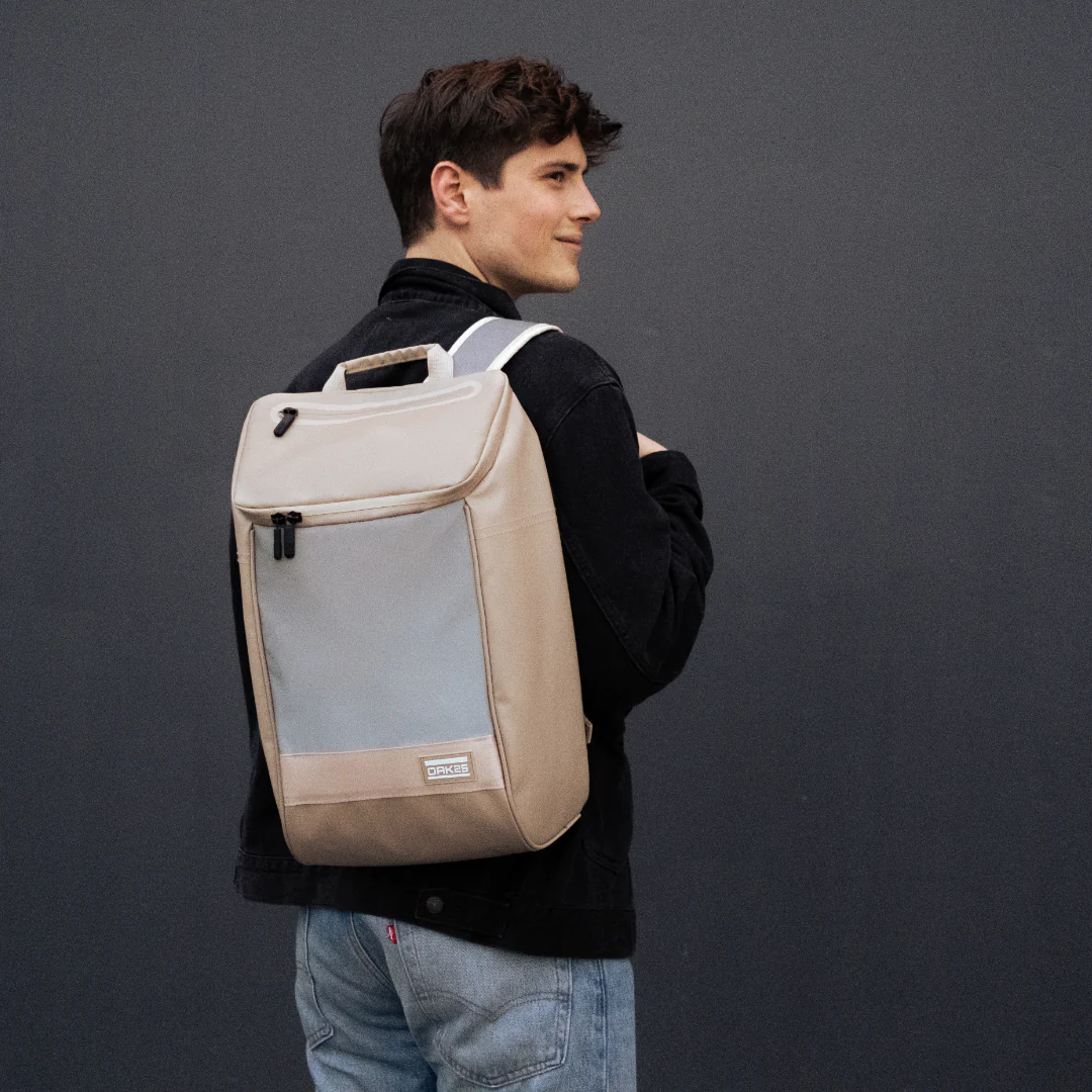 Mann trägt beigen Daybag-Rucksack mit reflektierendem Frontpanel und Label 'OAK25'