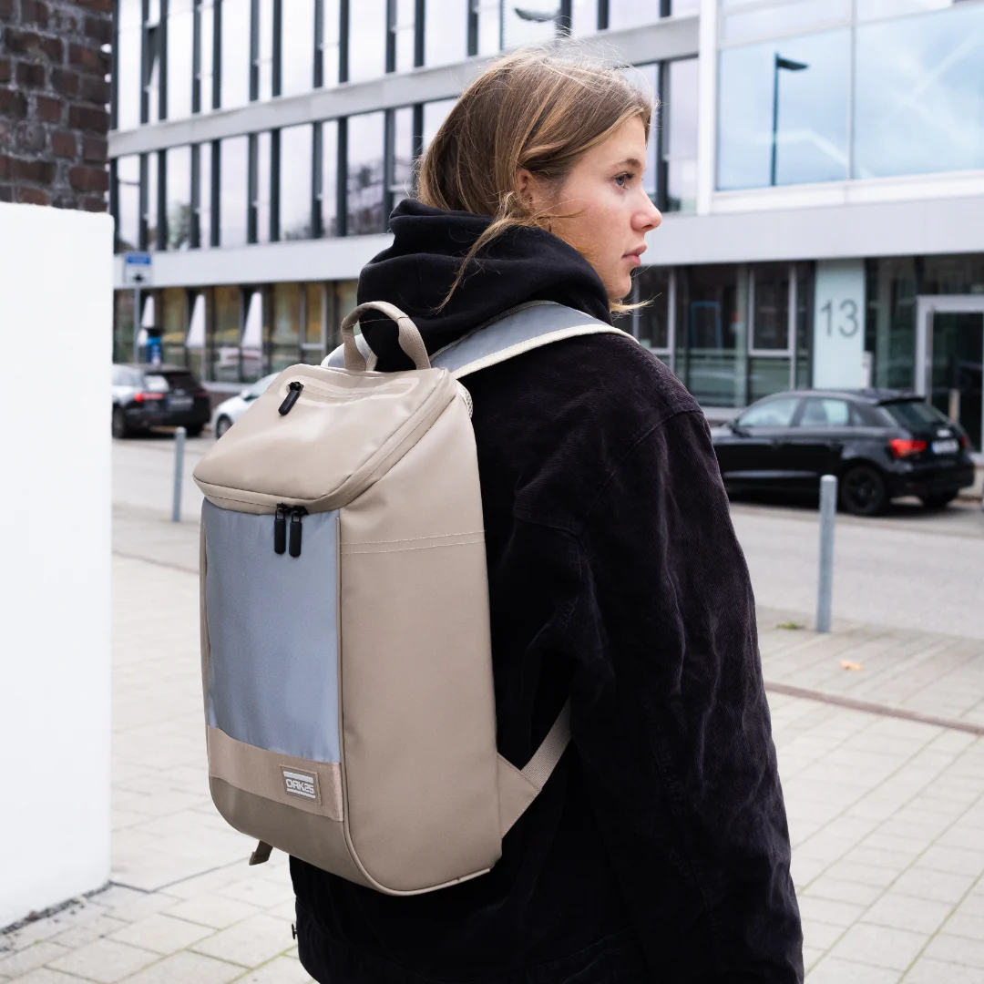 Frau trägt sandfarbenen, reflektierenden Rucksack mit zwei Reißverschlüssen vor einem modernen Bürogebäude