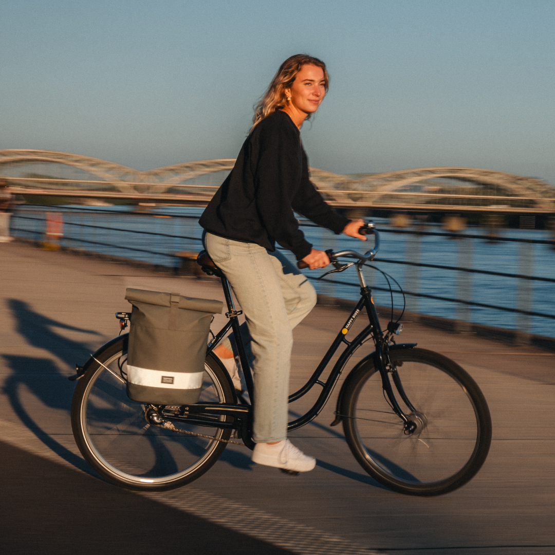 Frau fährt Fahrrad mit salbeigrünem rolltop Rucksack am Gepäckträger und reflektierendem Streifen an einem Wasserweg