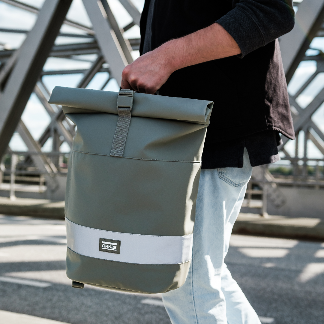 Person hält salbeifarbene rollbare Fahrradtasche mit reflektierendem Streifen und OAK25-Logo