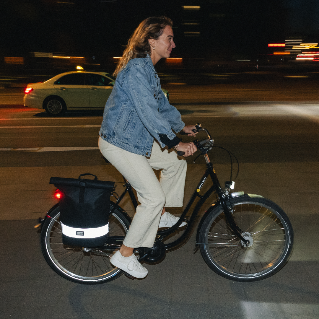 Frau mit Jeansjacke fährt nachts Fahrrad mit schwarzem, reflektierendem Rolltop-Rucksack auf Gepäckträger