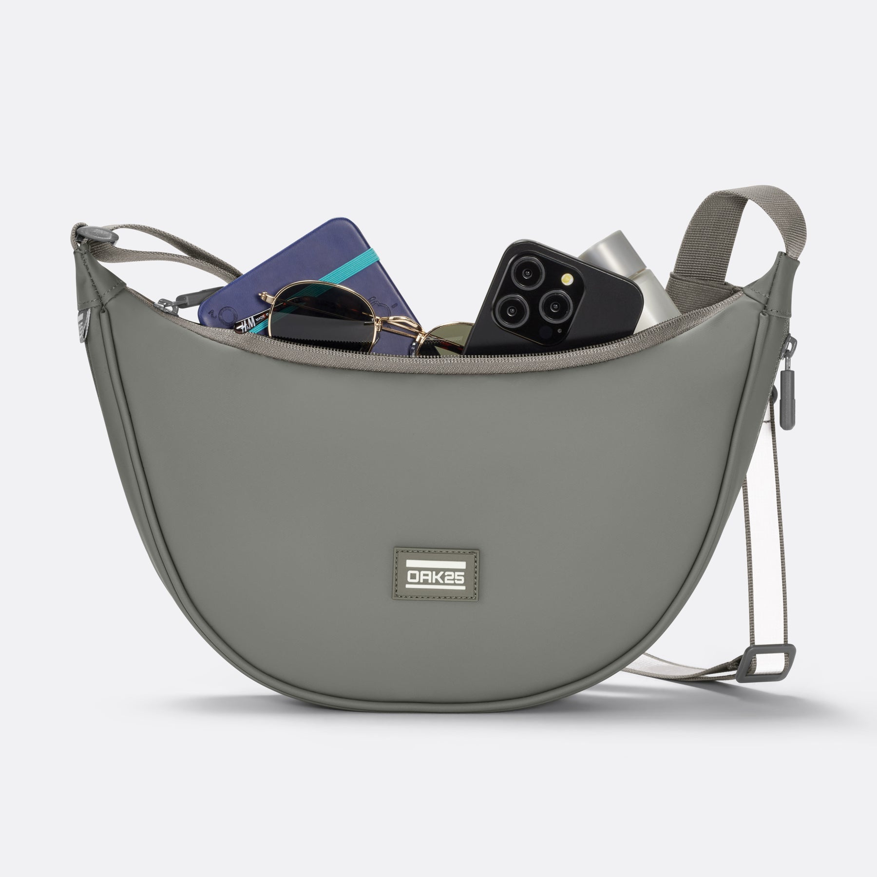 Salbeigrüne Half-Moon-Bag von OAK25 mit geöffnetem Hauptfach und sichtbar Sonnenbrille, Smartphone, Geldbeutel und Kosmetik