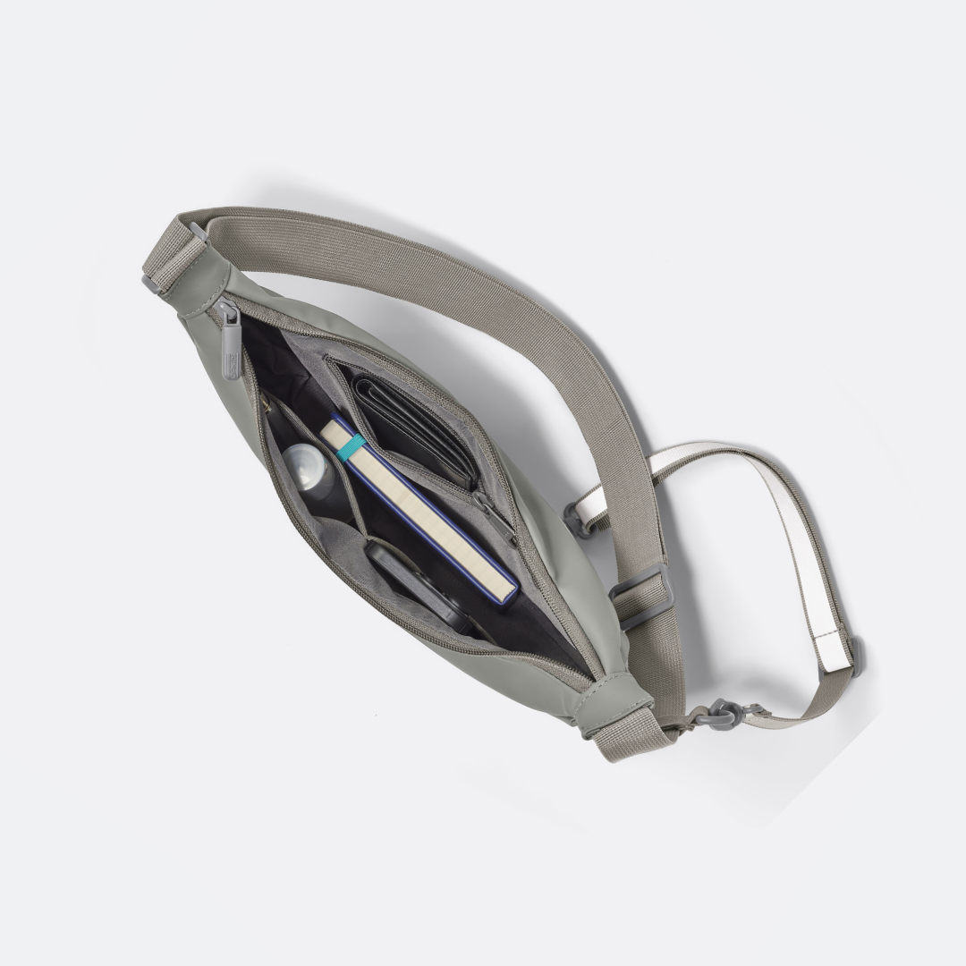 Salbeigrüne Halfmoonbag mit geöffnetem Hauptfach, gefüllt mit Geldbörse, Buch, Flasche und Handy