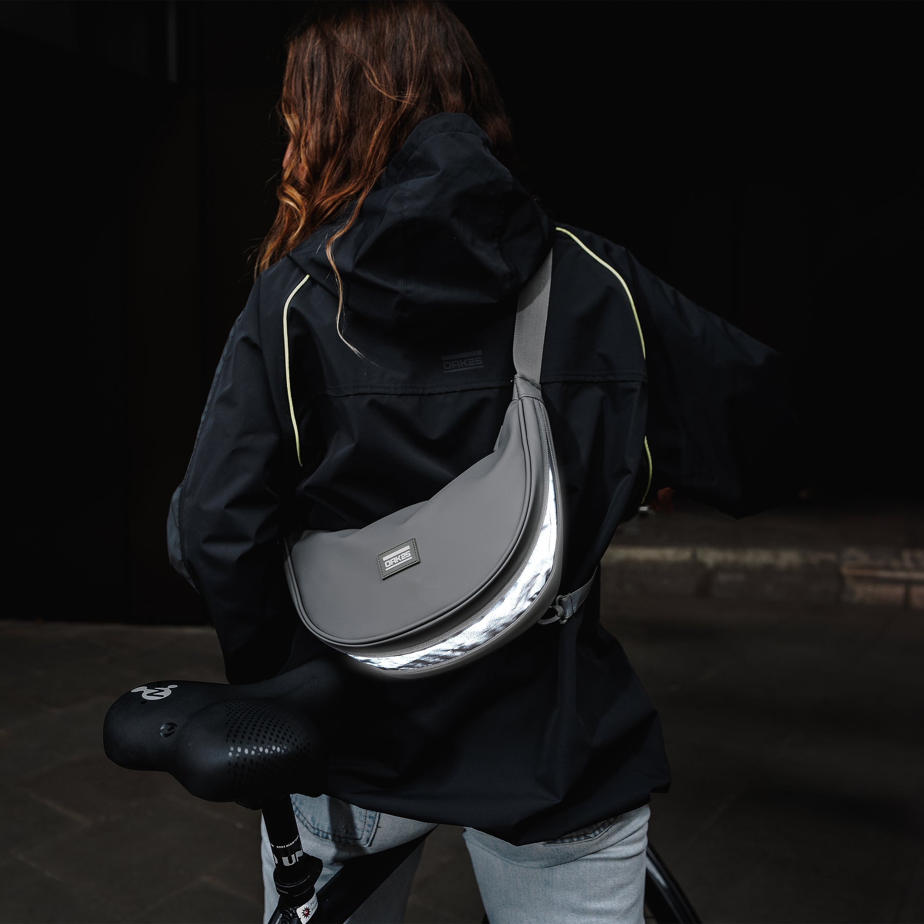 Rückenansicht einer Person mit schwarzer Jacke und grauer, halbmondförmiger Tasche mit reflektierendem Streifen