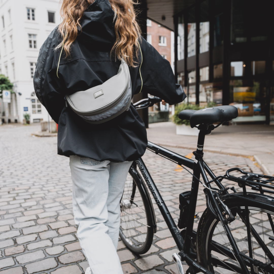 Person mit schwarzer Jacke und hellgrauer Halfmoon-Tasche zieht ein schwarzes Fahrrad auf gepflastertem Weg