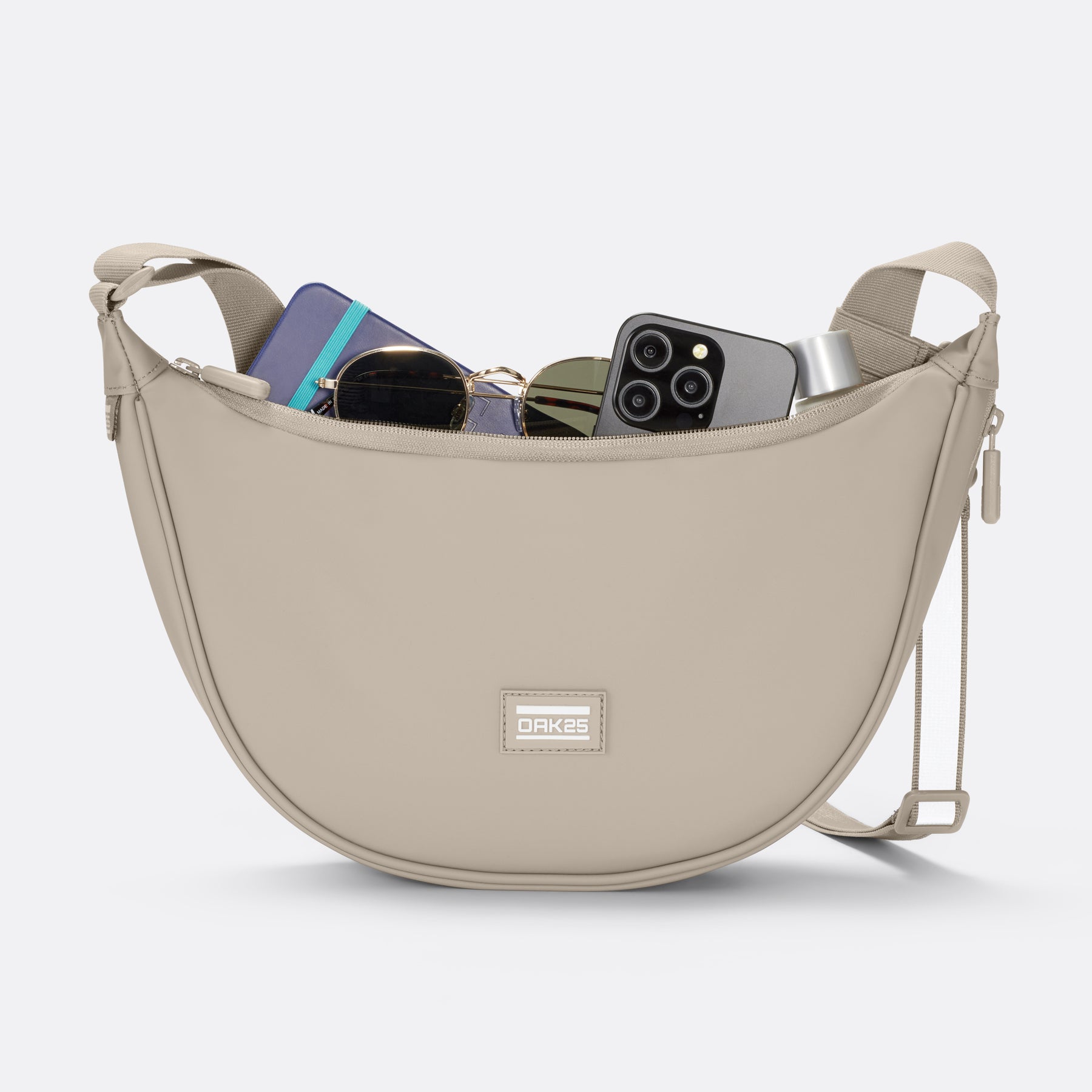 Sandfarbene Halbmond-Tasche von OAK25 mit Reißverschluss geöffnet, zeigt Sonnenbrille, Handy, Geldbörse und Lippenstift im Innenraum