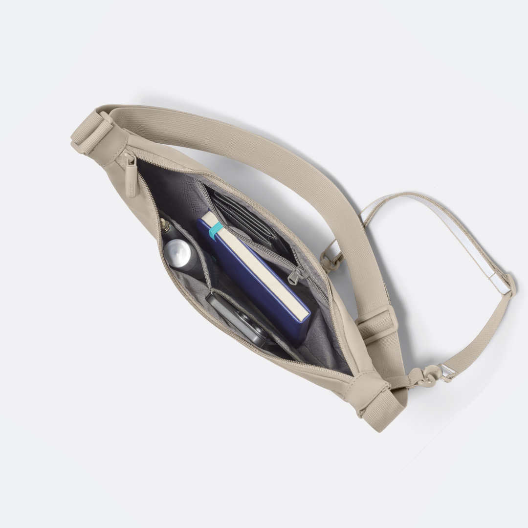 Sandfarbene Halbmondtasche mit Reißverschluss, geöffnet mit Buch, Flasche und Handy innen