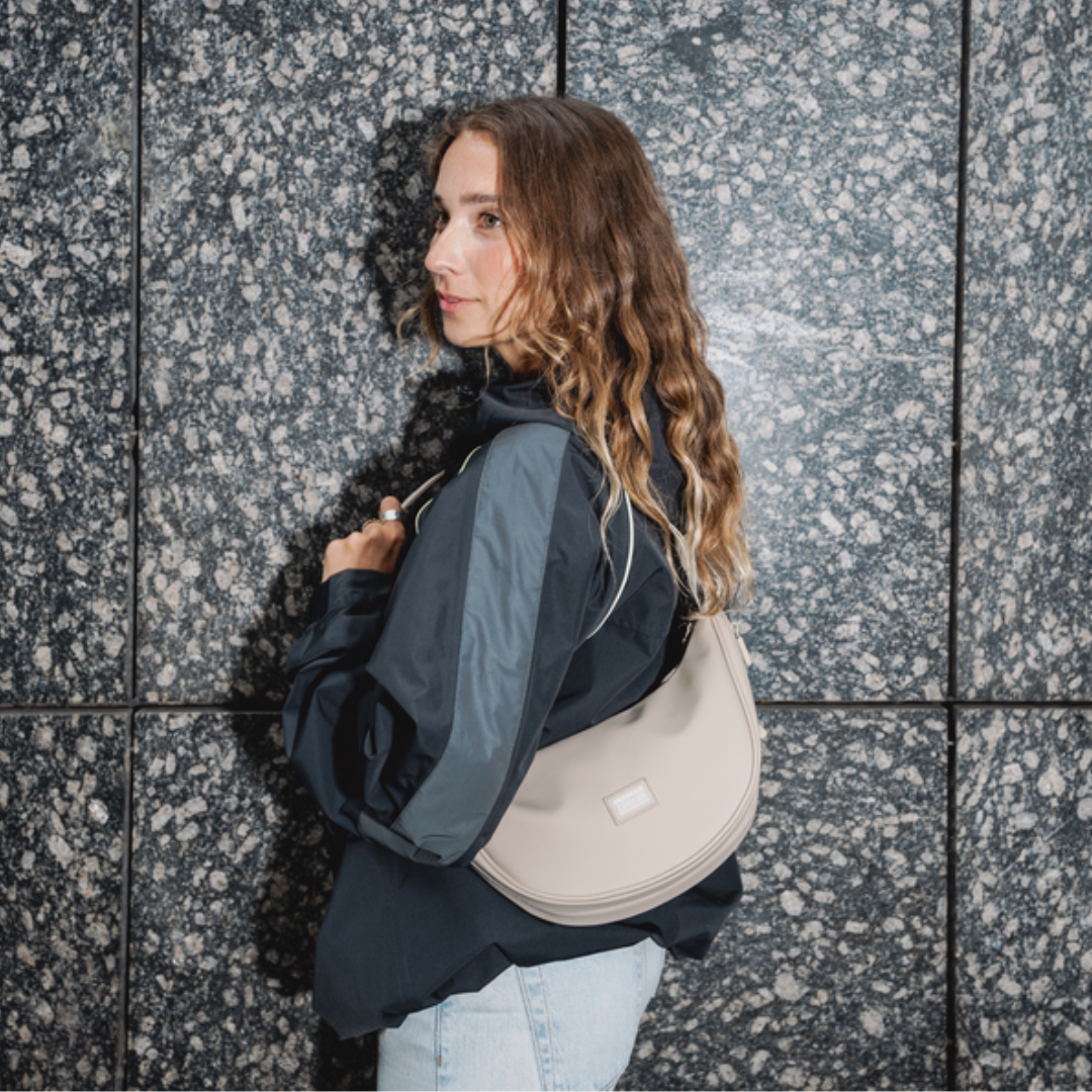 Frau trägt eine sandfarbene Halbmondtasche der Marke OAK25 über der Schulter vor einer grauen Steinwand