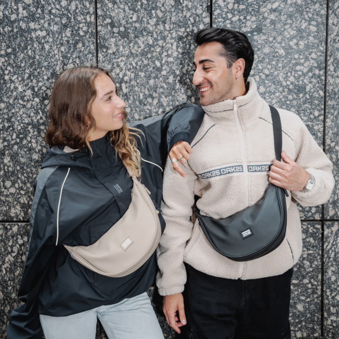 Zwei Personen tragen halbmondförmige Umhängetaschen, eine beige und eine schwarze, vor einer grauen Steinwand