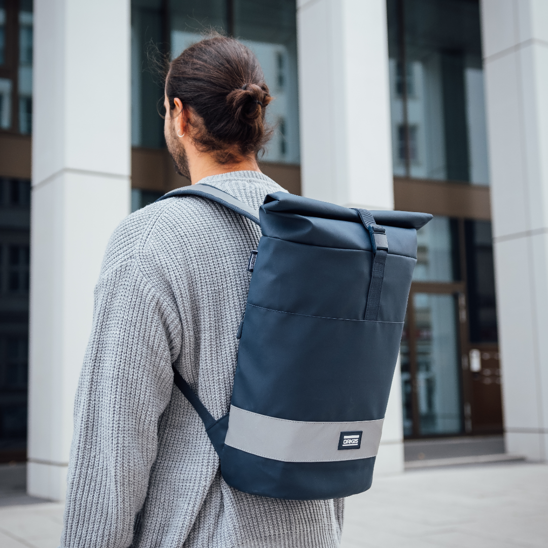 Rucksack OAK25 Everyday Rolltop in Navy mit reflektierendem Streifen am Gepäckträger getragen von Person mit grauem Pullover