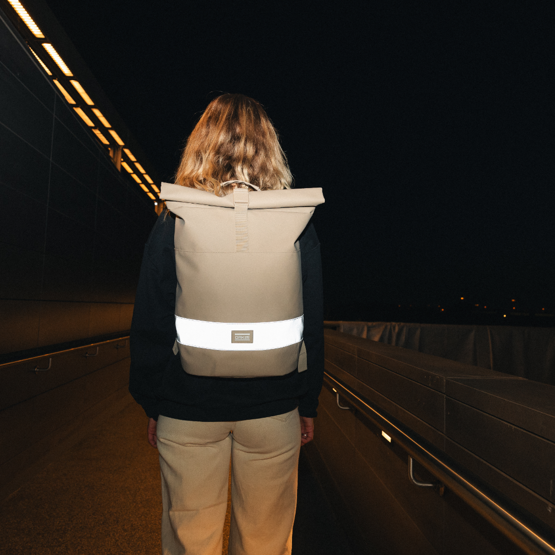 Person von hinten mit sandfarbenem OAK25 Everyday Rolltop Rucksack mit reflektierendem Streifen auf Gepäckträger bei Nacht