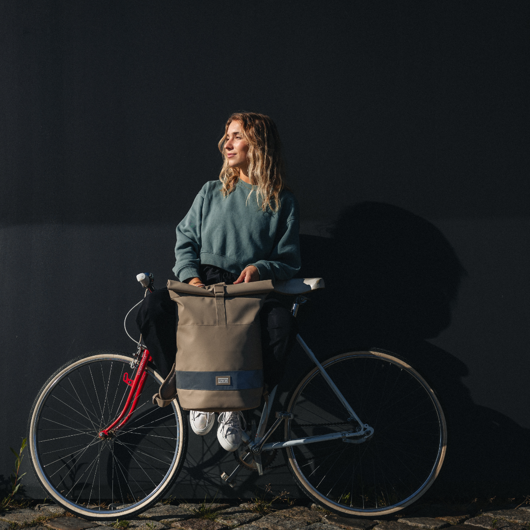 Frau mit sandfarbener, reflektierender Rolltop-Fahrradtasche sitzt auf einem Fahrrad vor schwarzer Wand