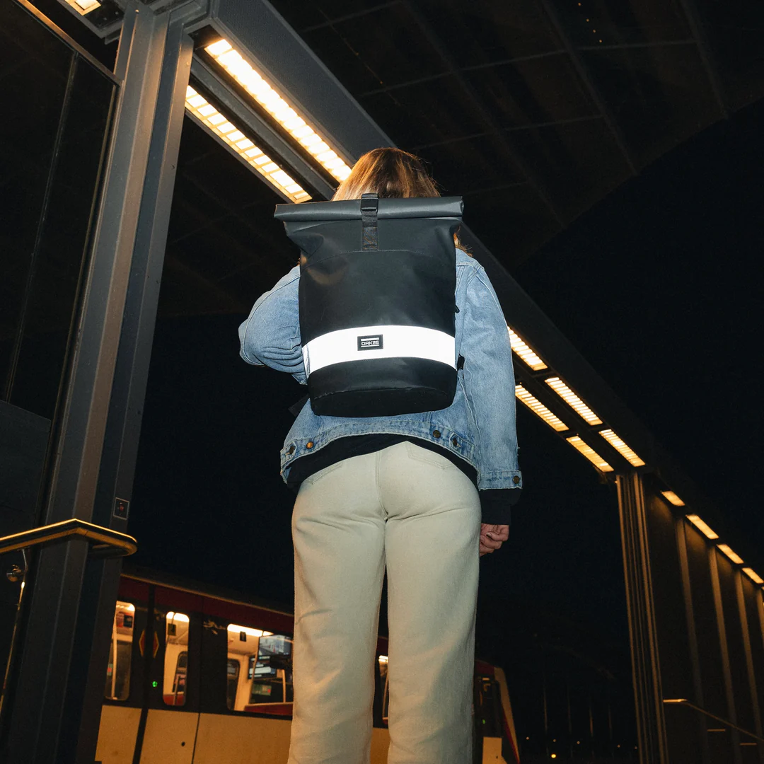 Person von hinten trägt schwarzen rolltop Rucksack mit reflektierendem Streifen und ORK logo, trägt Jeansjacke und helle Hose