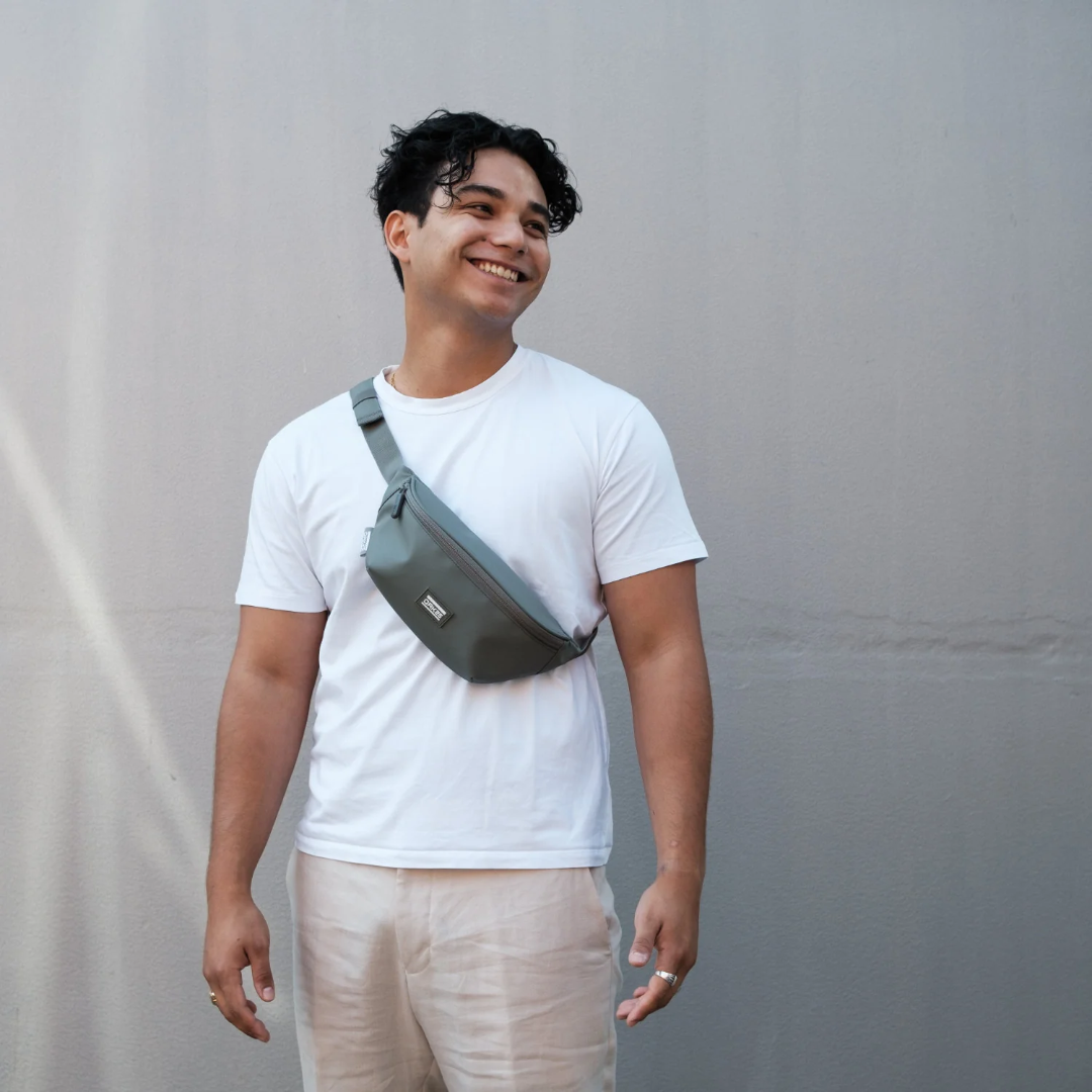 Mann mit kurzen lockigen Haaren trägt eine salbeigrüne Bauchtasche quer über der Brust und ein weißes T-Shirt