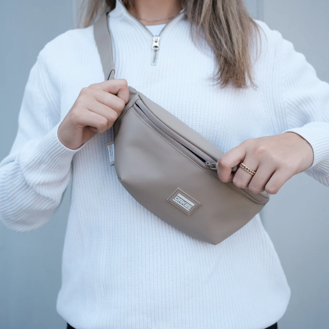 Person trägt eine sandfarbene OAKS Bauchtasche über der Schulter und öffnet den Reißverschluss