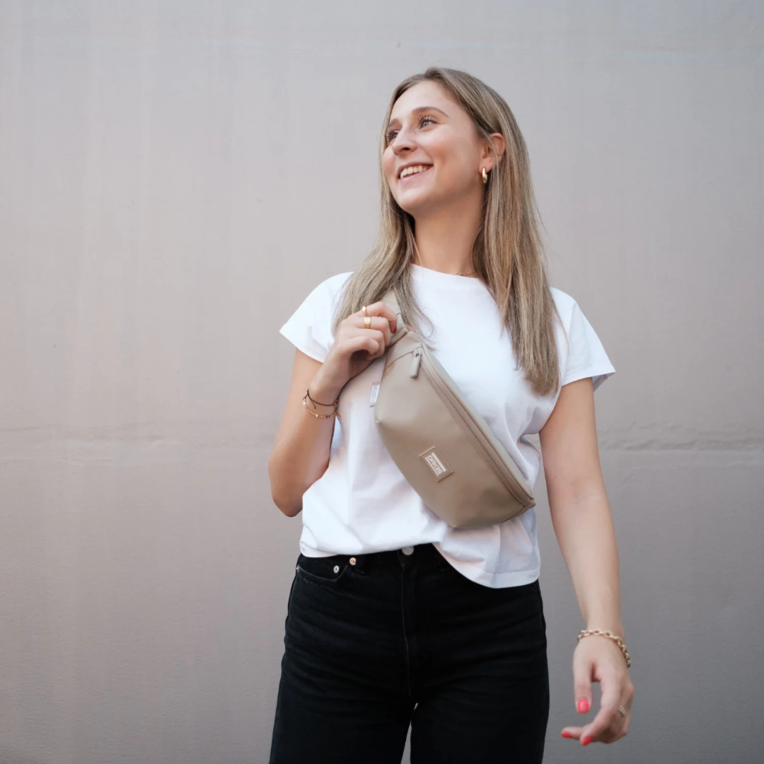 Frau trägt sandfarbene Bauchtasche über weiße T-Shirt und schwarze Hose, lächelnd nach rechts blickend