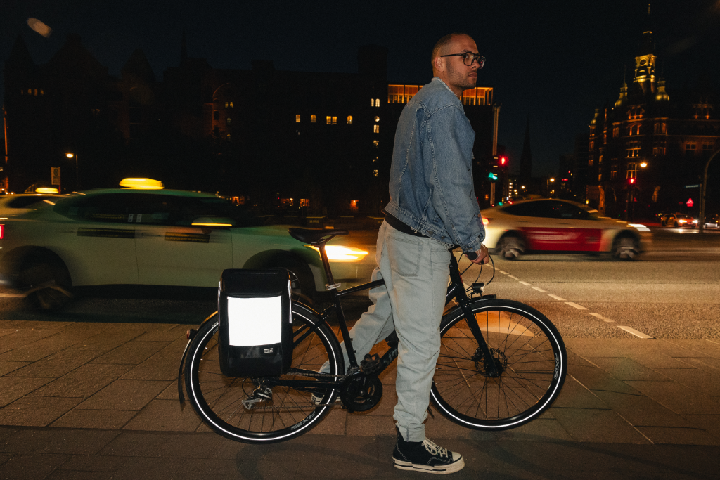 Mann mit Jeansjacke steht mit Fahrrad bei Nacht, reflektierende Fahrradtasche am Hinterrad