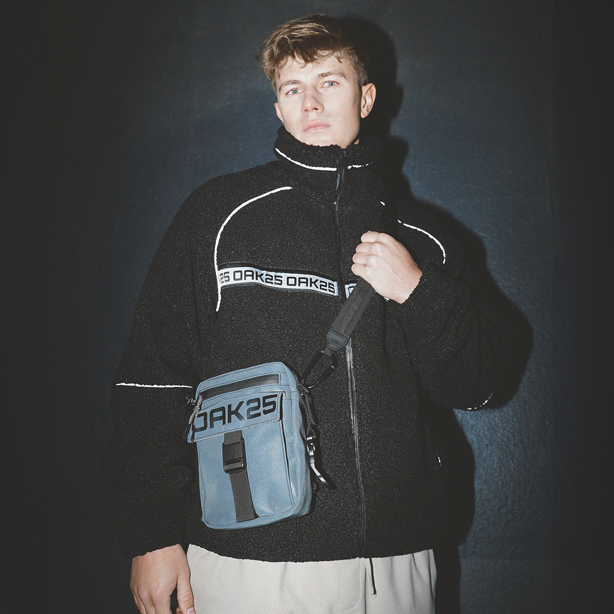 Junge trägt schwarze Fleecejacke mit OAK25 Schriftzug und blaue, wetterfeste Bauchtasche vor der Brust