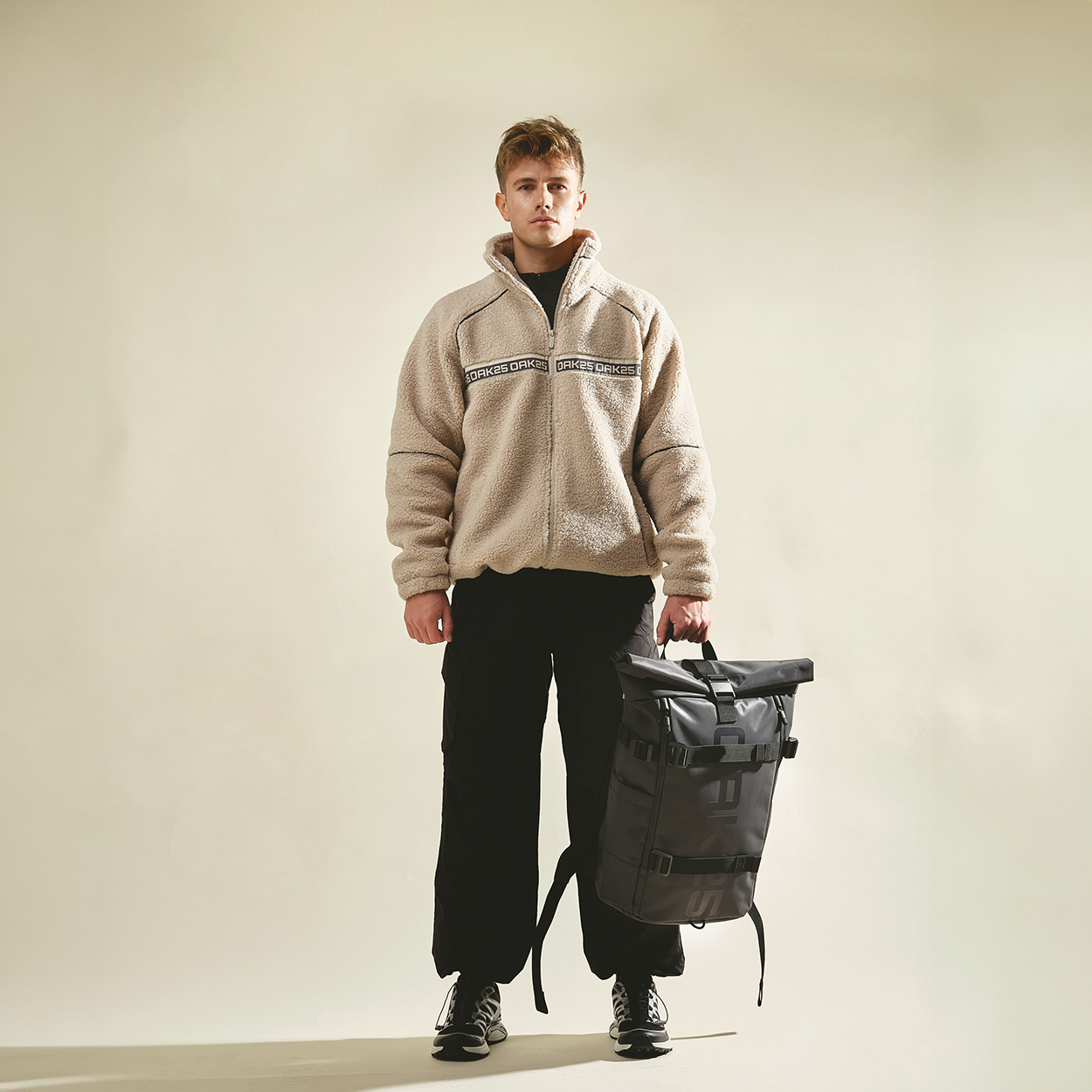 Mann trägt beige Teddyjacke und schwarze Hose, hält schwarzen, reflektierenden Rolltop-Rucksack von OAK25 in der Hand