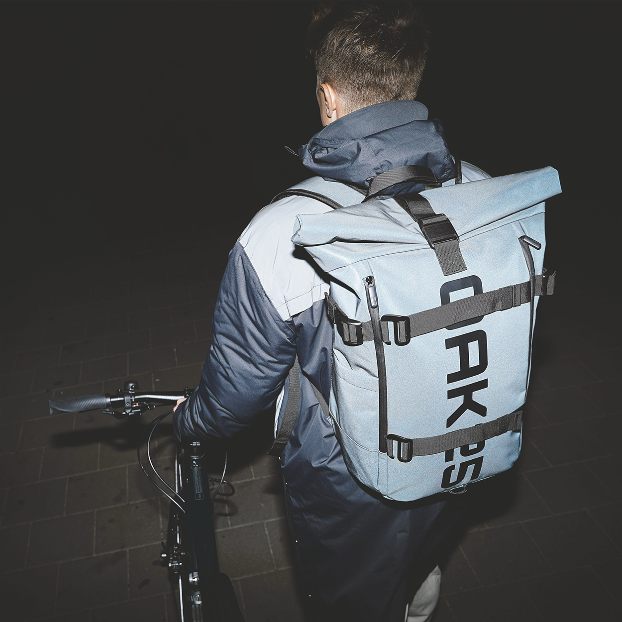 Person von hinten mit schwarzem Fahrrad und hellblauem OAK25 Luminite Rolltop Rucksack