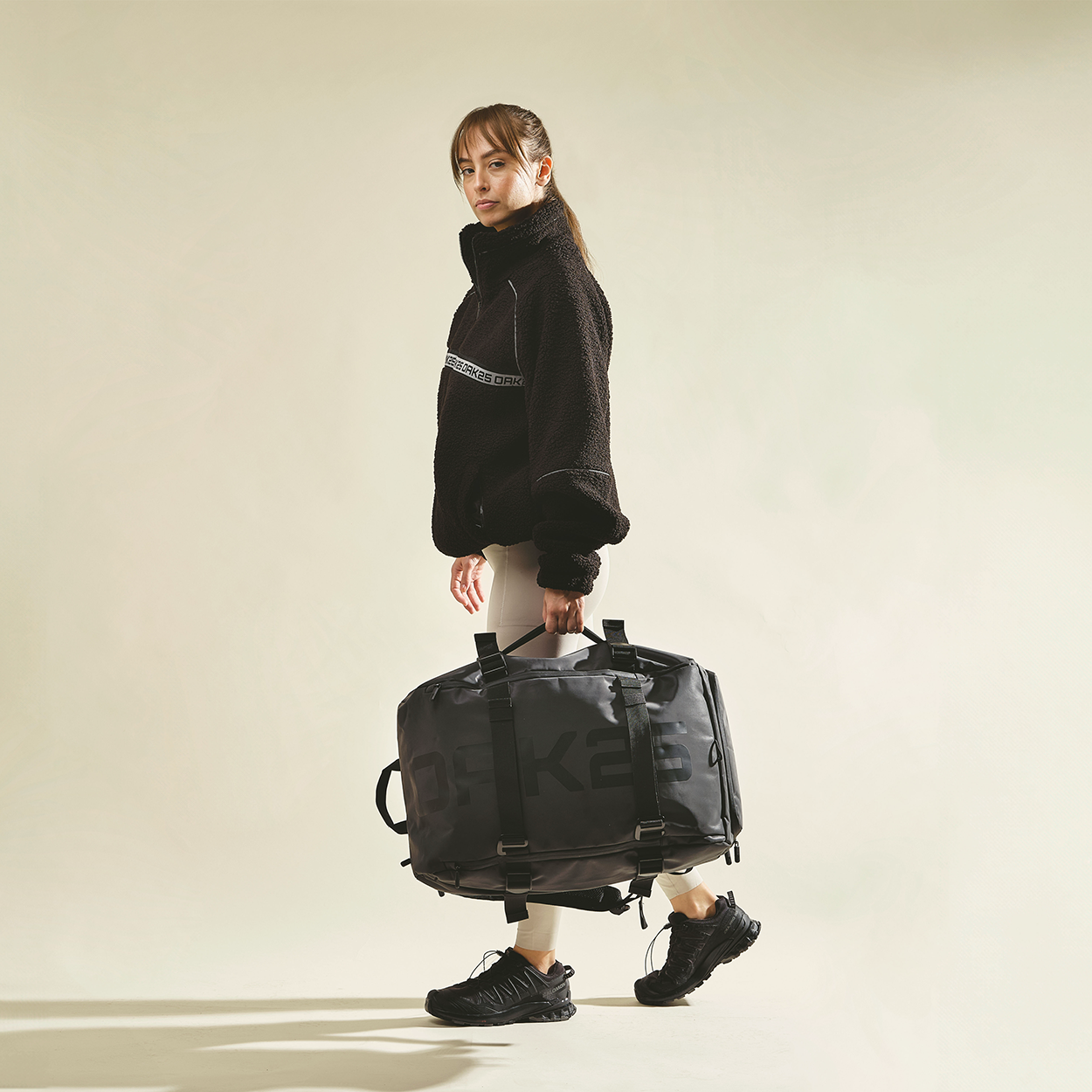Frau in schwarzem Pullover und beige Hose trägt große schwarze Dufflebag mit OAK25-Logo
