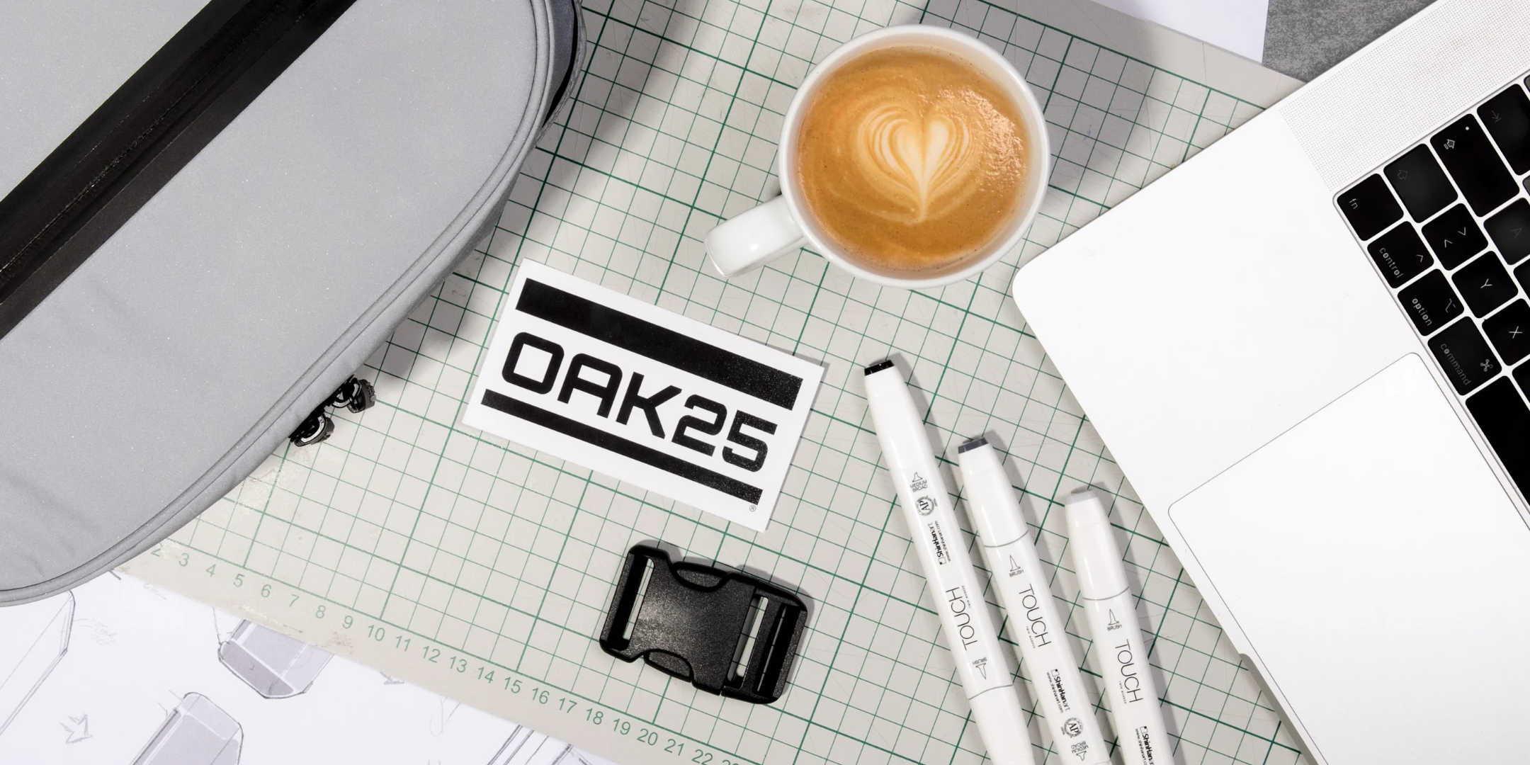 Arbeitsplatz mit Laptop, Kaffeetasse mit Herzmilchschaum, drei Marker, OAK25-Aufkleber, Gurtverschluss und grauer Tasche auf kariertem Schneidbrett