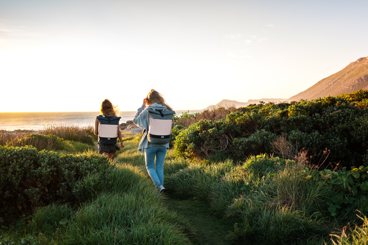 Zwei Personen wandern mit Rolltop-Rucksäcken aus recyceltem PET entlang eines Küstenpfads bei Sonnenuntergang