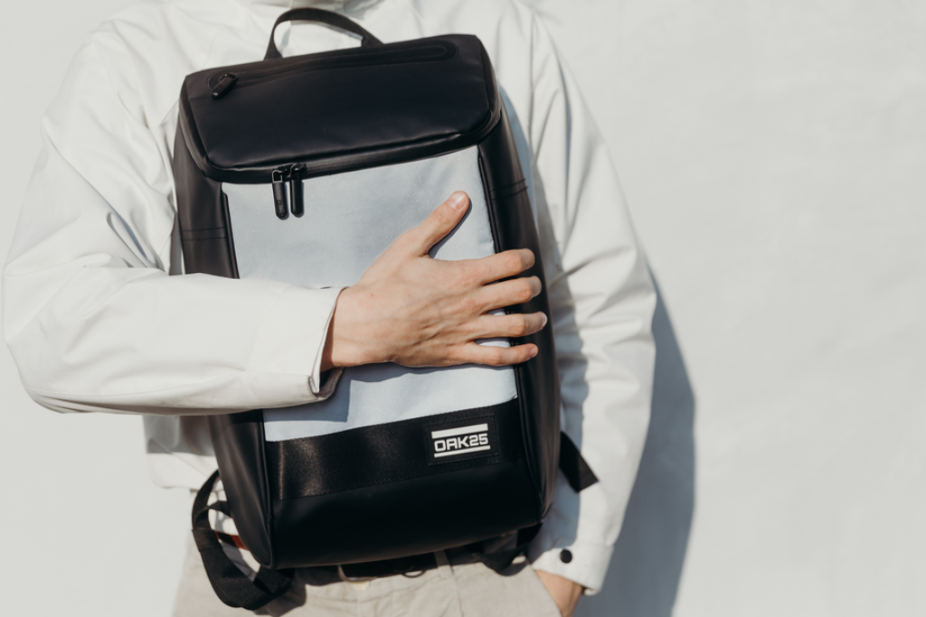 Person trägt weißen Mantel und hält einen zweifarbigen Rucksack mit OAK25-Logo