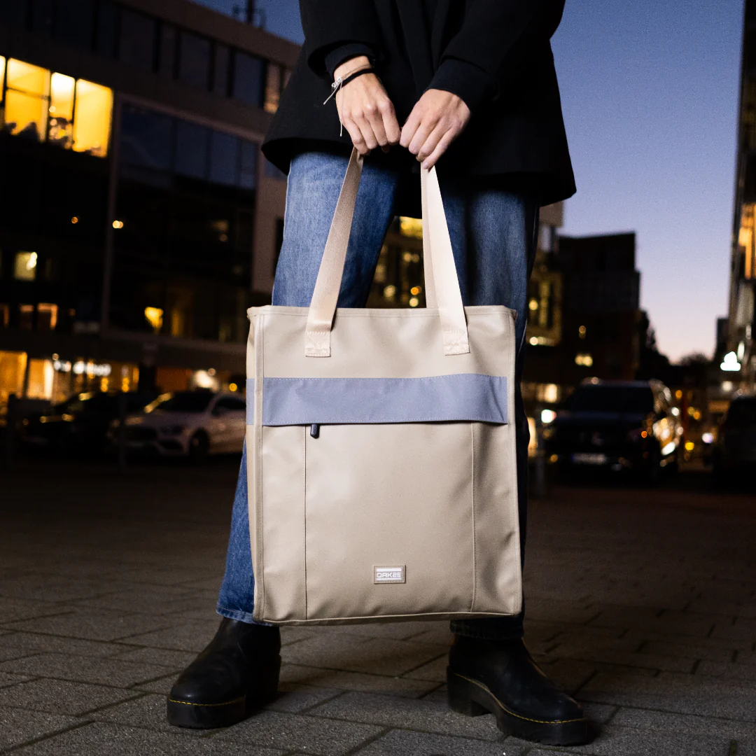 Sandfarbener reflektierender Totebag von DAKKS, getragen von einer Person in Jeans und schwarzen Stiefeln