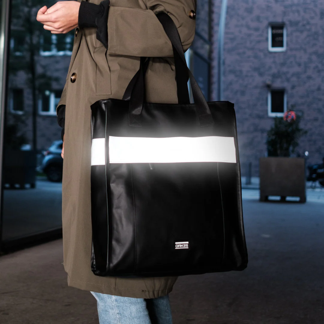 Schwarze reflektierende Totebag mit weißem Streifen und OAK25-Logo, getragen von Person in beigefarbenem Mantel