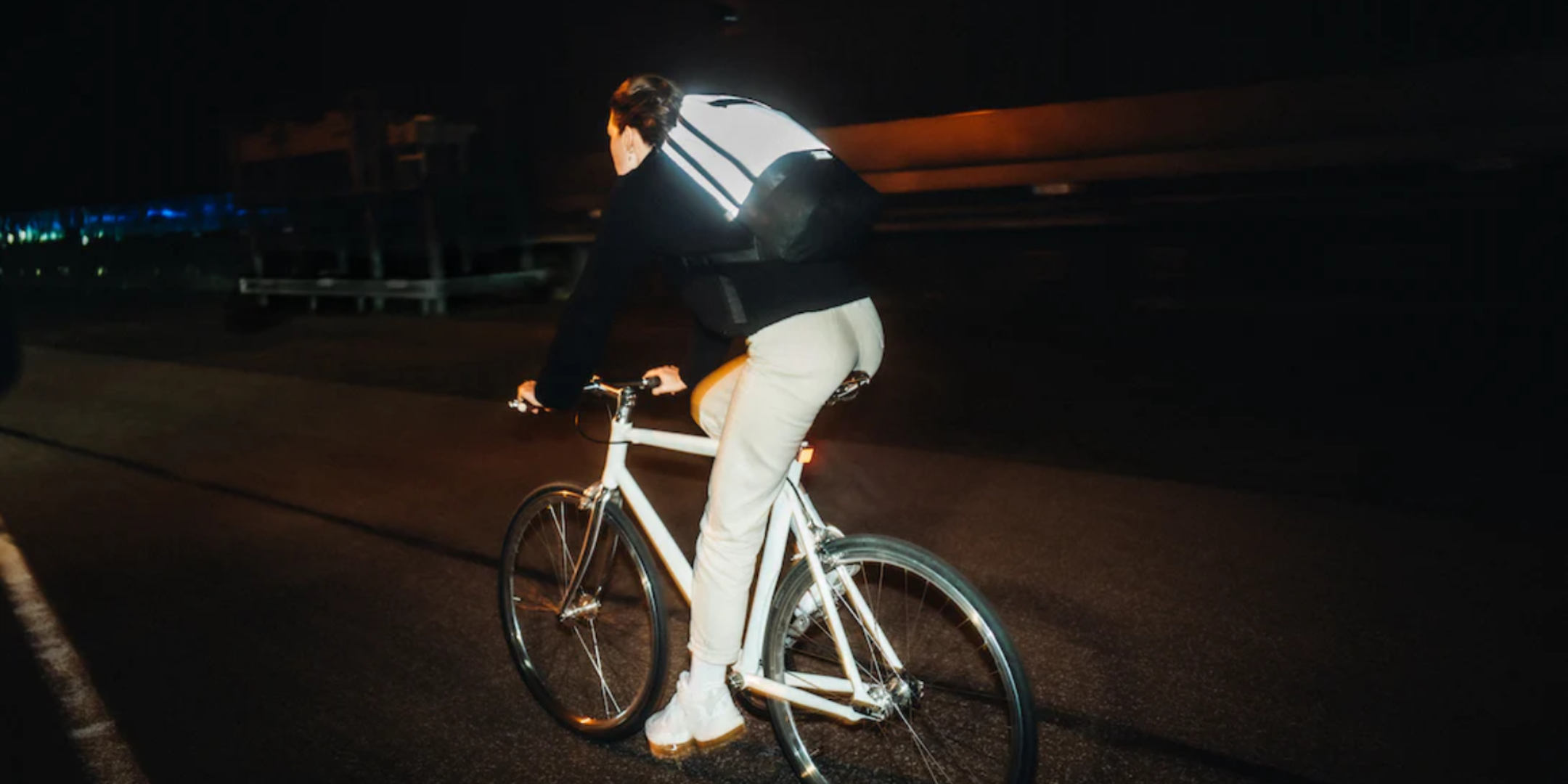 Person in heller Hose und dunklem Pullover fährt nachts Fahrrad mit reflektierendem Rucksack