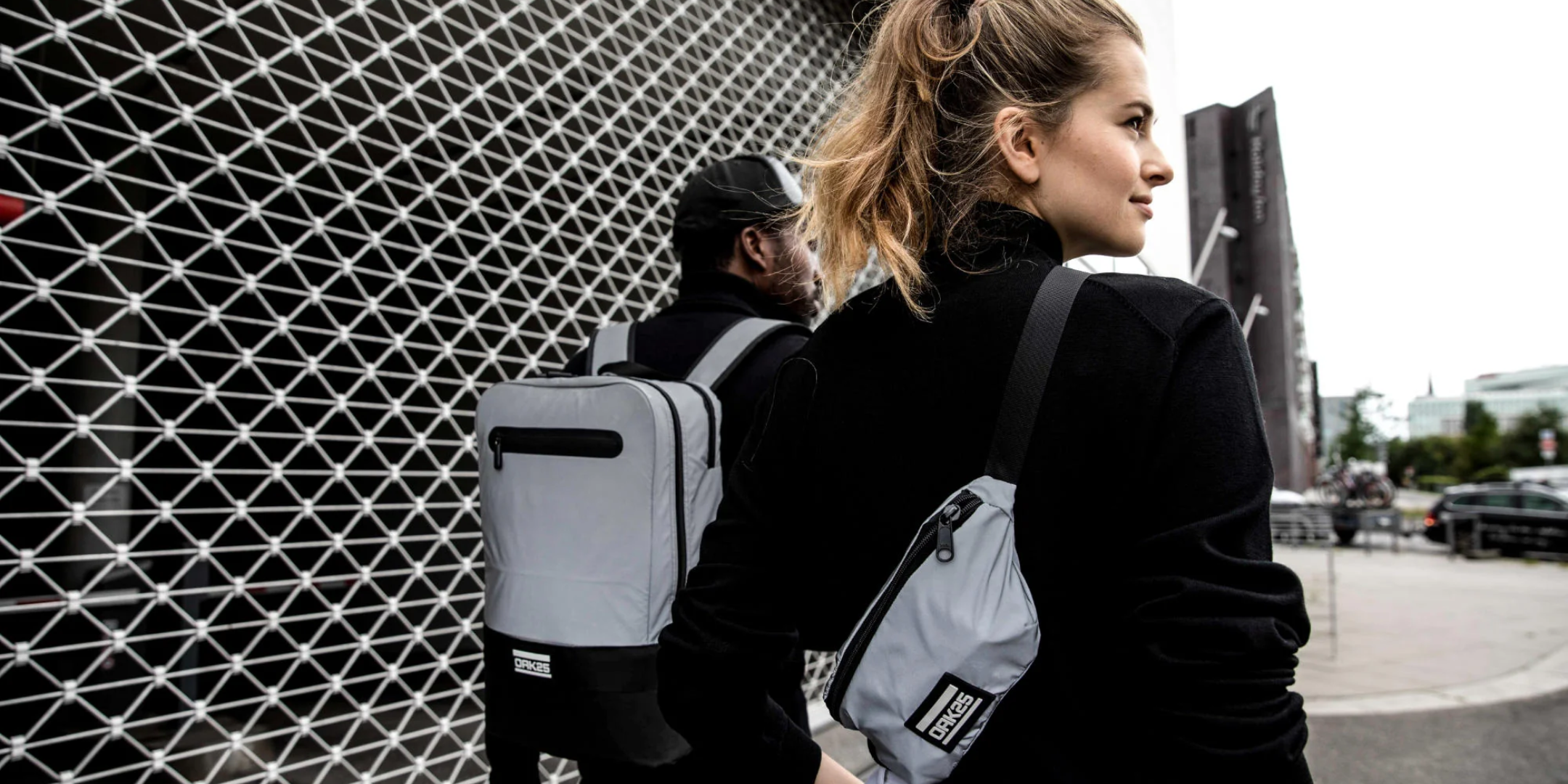 Zwei Personen tragen reflektierende Taschen von OAK25, eine als Rucksack, eine als Umhängetasche, vor einer urbanen Wand