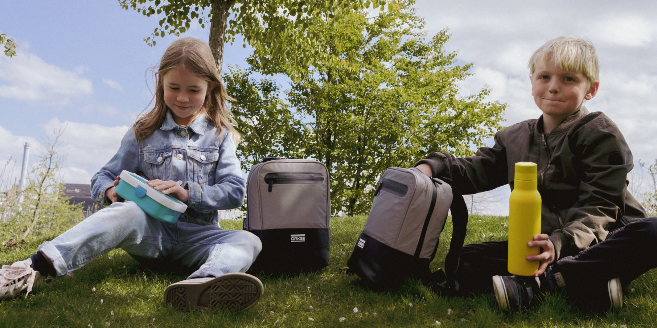 Mädchen und Junge sitzen auf einer Wiese mit nachhaltigen Rucksäcken, das Mädchen hält eine Brotdose, der Junge eine gelbe Trinkflasche