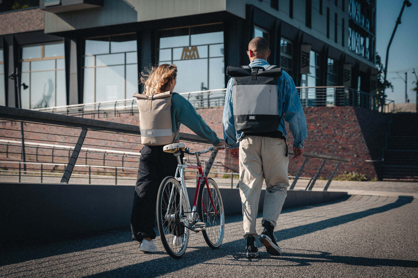 Junge Frau mit hellbraunem reflektierendem Rucksack schiebt Fahrrad, junger Mann mit schwarz-grauem reflektierendem Rucksack geht neben ihr auf Promenade